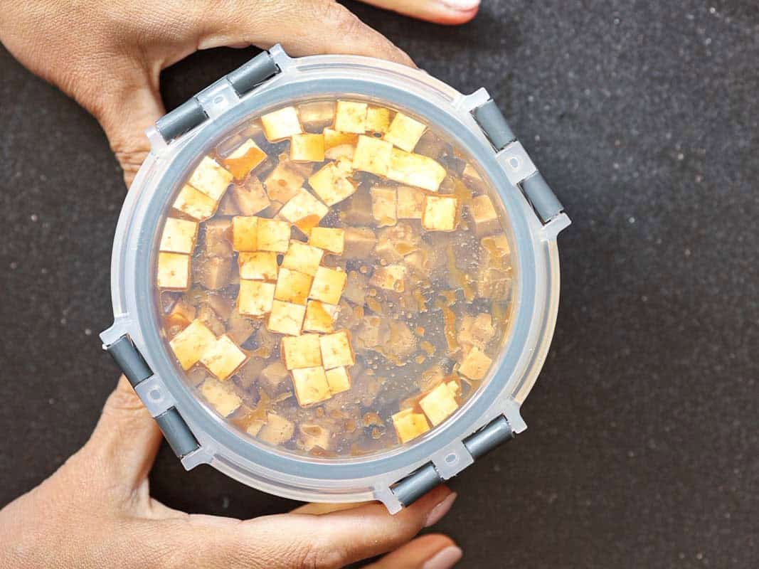 Hands holding a round, transparent container filled with small, diced pieces of tofu submerged in a liquid marinade. The container rests on a dark surface.